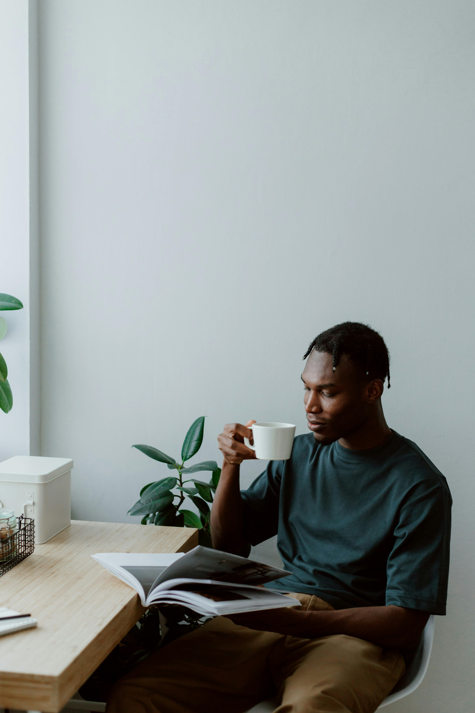 A young man sitting at a light wood desk drinking from a white mug while reading a book