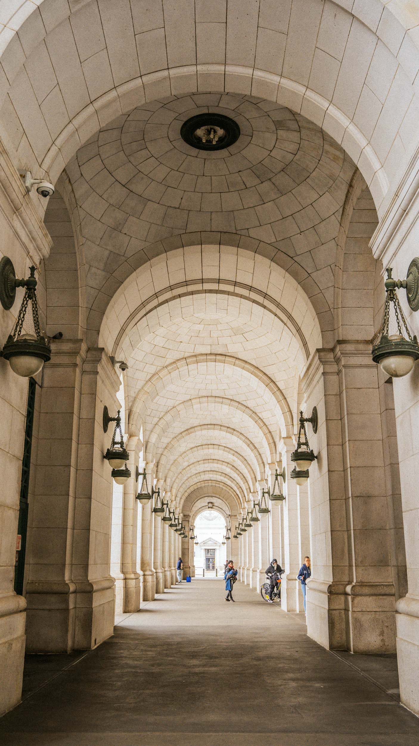 Perspective view of a long vaulted stone corridor with classical arches and hanging lanterns